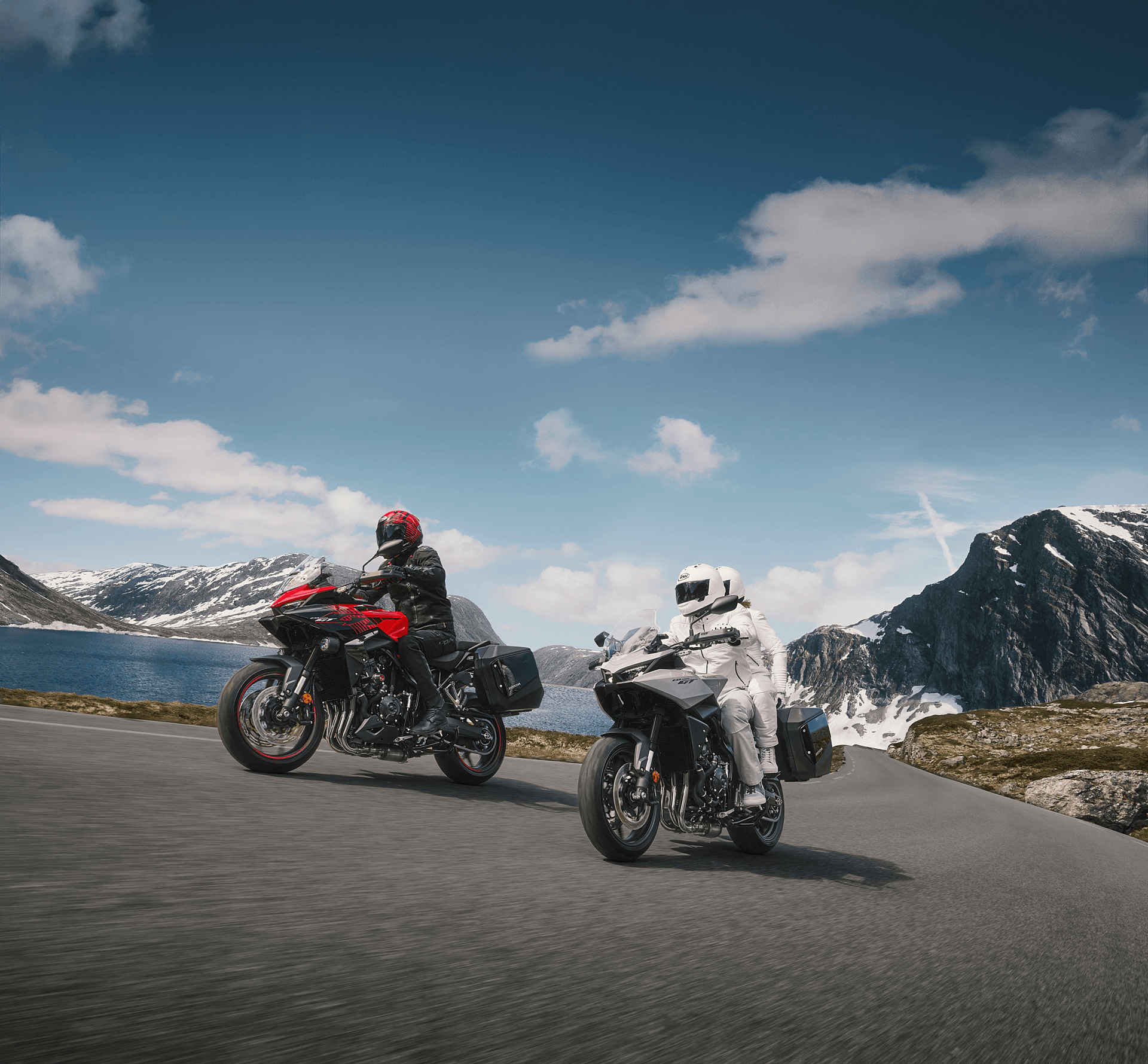 Two motorcyclists on a mountain road with a lake and snow-capped peaks in the background.