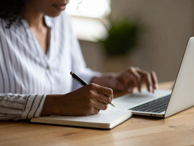 Person writing in a notebook and typing on a laptop at a wooden desk.