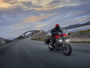 Motorcyclist in black gear and red helmet riding on a curved open road at dusk.