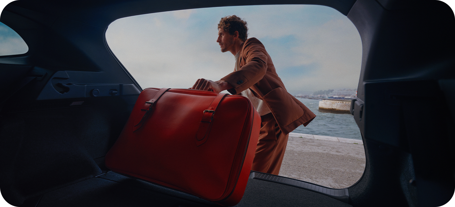 Man in a suit placing a red leather bag into an open car boot by the water.