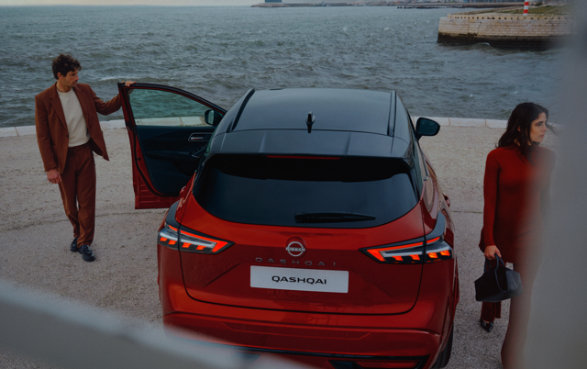 Rear view of a red Nissan Qashqai parked by the sea, with two people nearby.