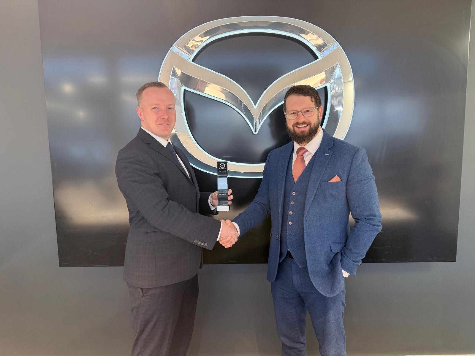 Two men in suits shaking hands in front of a Mazda logo, one holding an award.