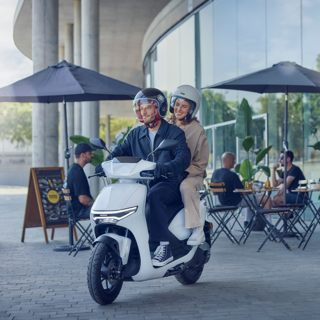 Two people on a white scooter wearing helmets, riding past an outdoor café.