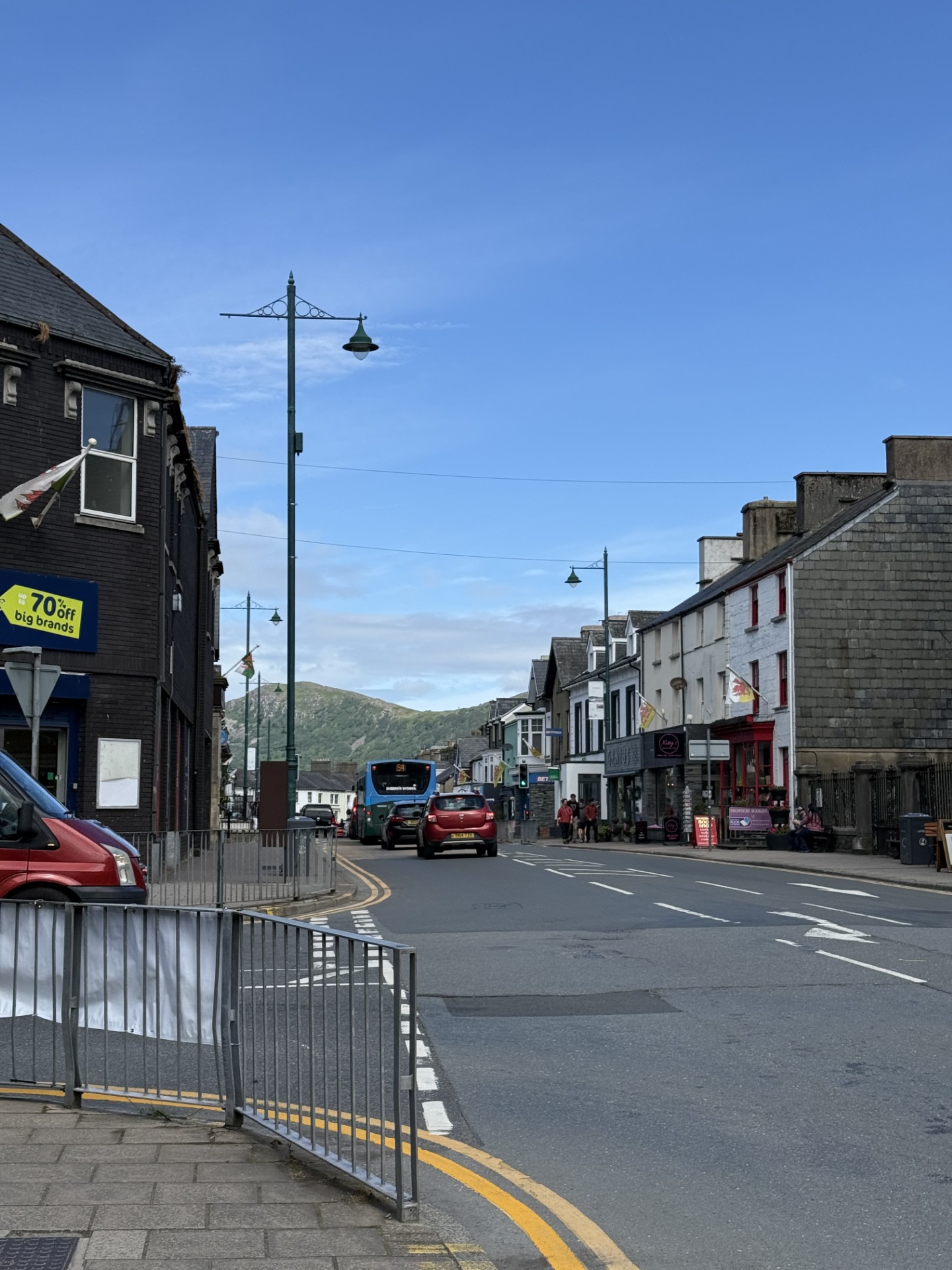 A street view with shops, cars, and a bus in a small town. Hills are visible in the distance.