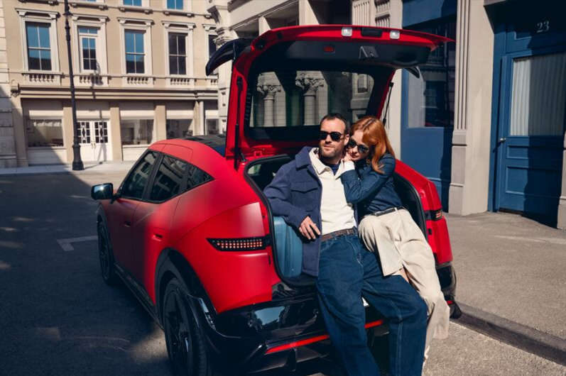 Couple sitting in the boot of a red car, with its hatch open in an urban setting.