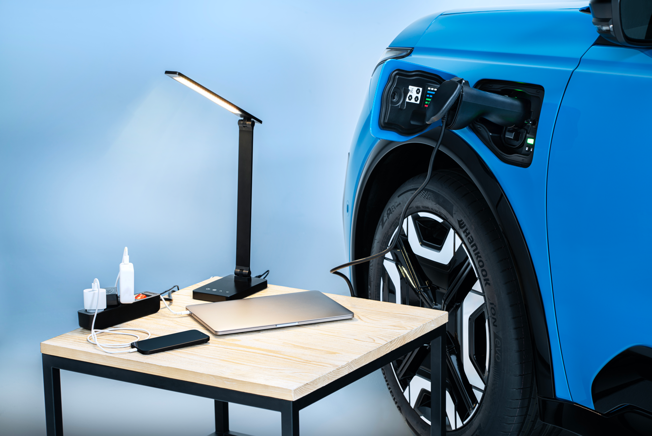 Electric car charging with a table holding a lamp, laptop, phone, and charging accessories.