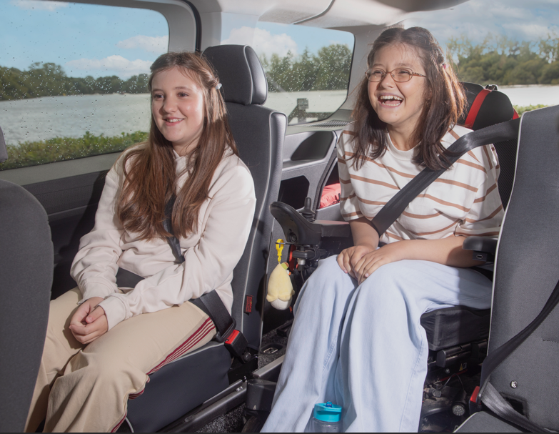 Two girls sitting in a car, both wearing seatbelts and smiling.
