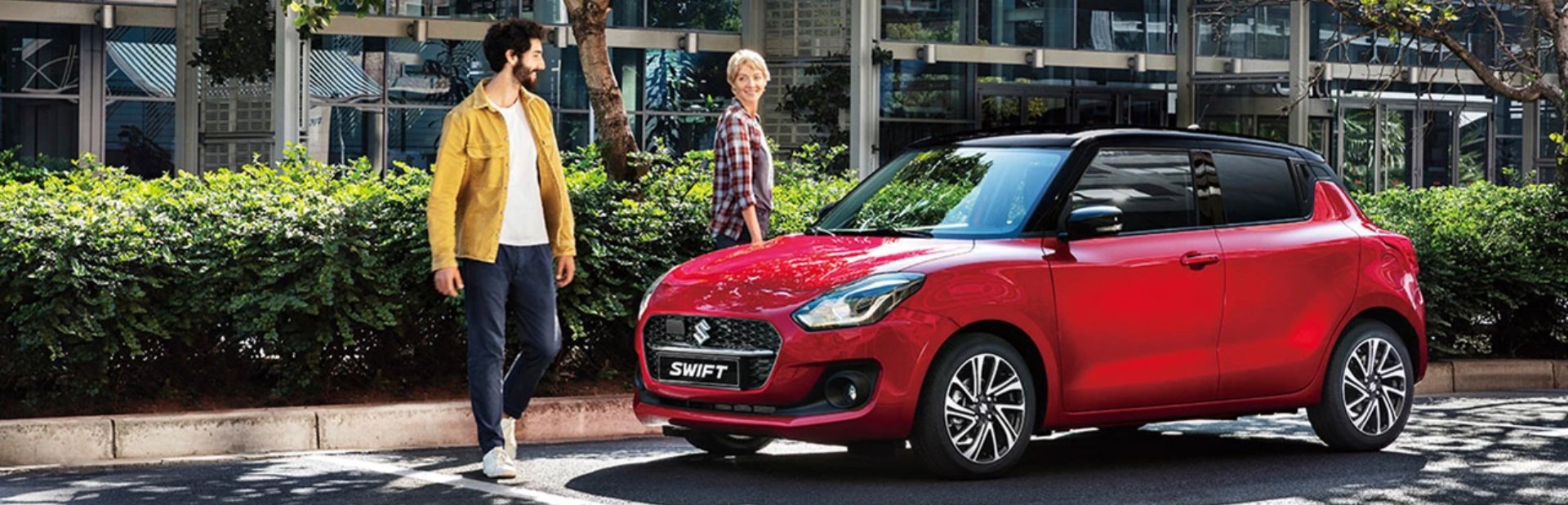 Two people walking near a red hatchback car.